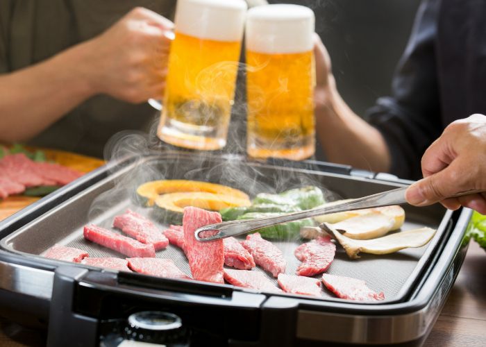 Two people clink mugs of beer as someone grills wagyu beef in the foreground